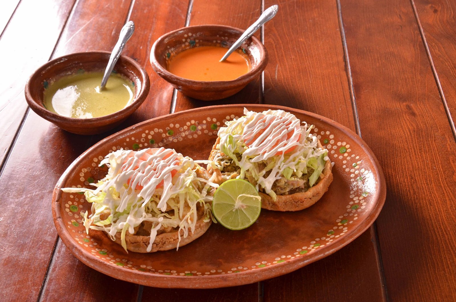 Tasty Mexican sopes topped with lettuce, sour cream, and served with lime and sauces on a wooden table.